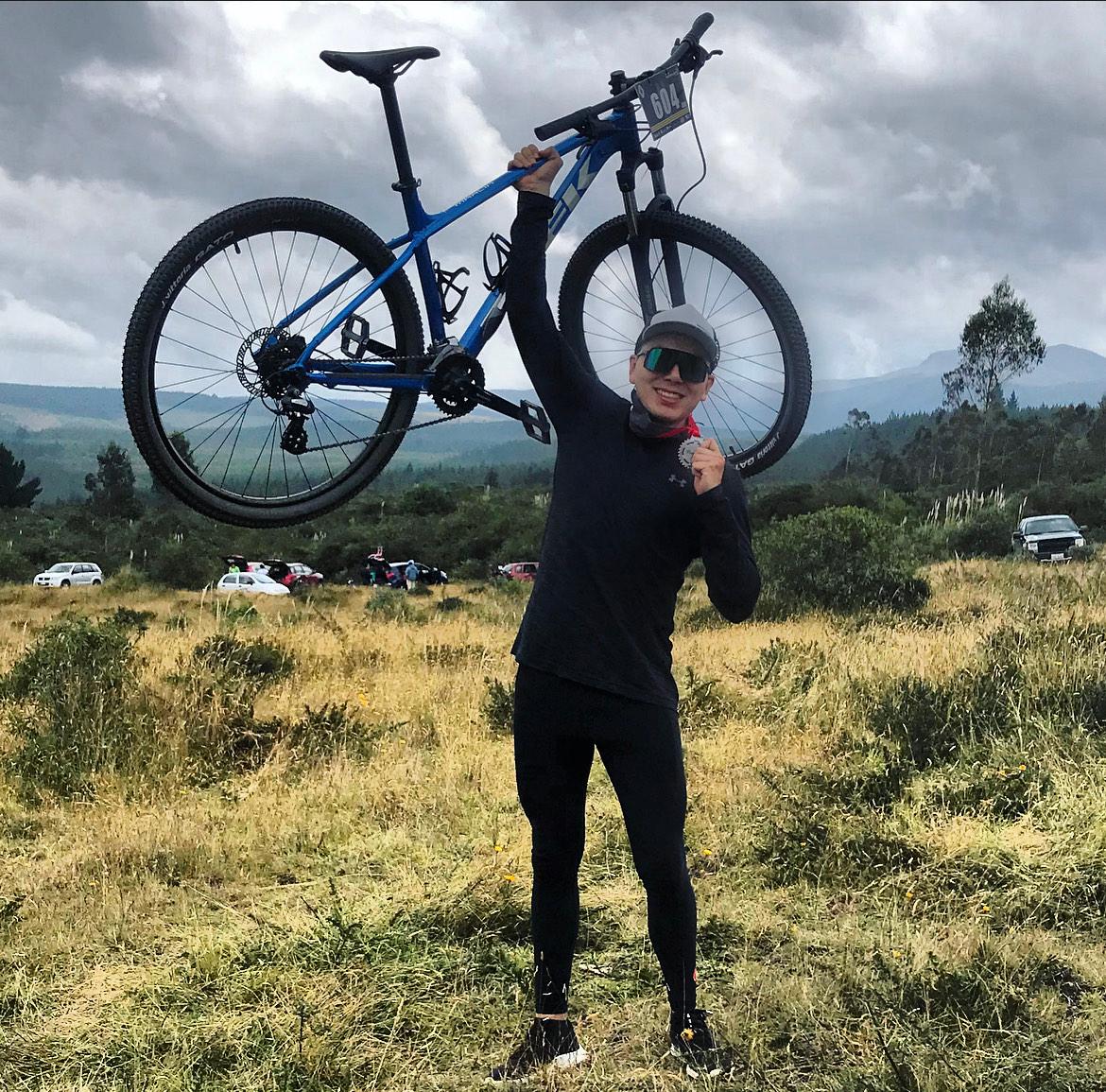 Jorge levanta su bicicleta de montaña sobre la cabeza, con una medalla en la mano, en un paisaje andino
